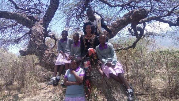 Rescued Kenyan Masaii girls in purple and grey school uniform sit in a tree under a blue sky. In their midst sits the matron from their safe house , Beatrice, and the charity founder, Jonathan